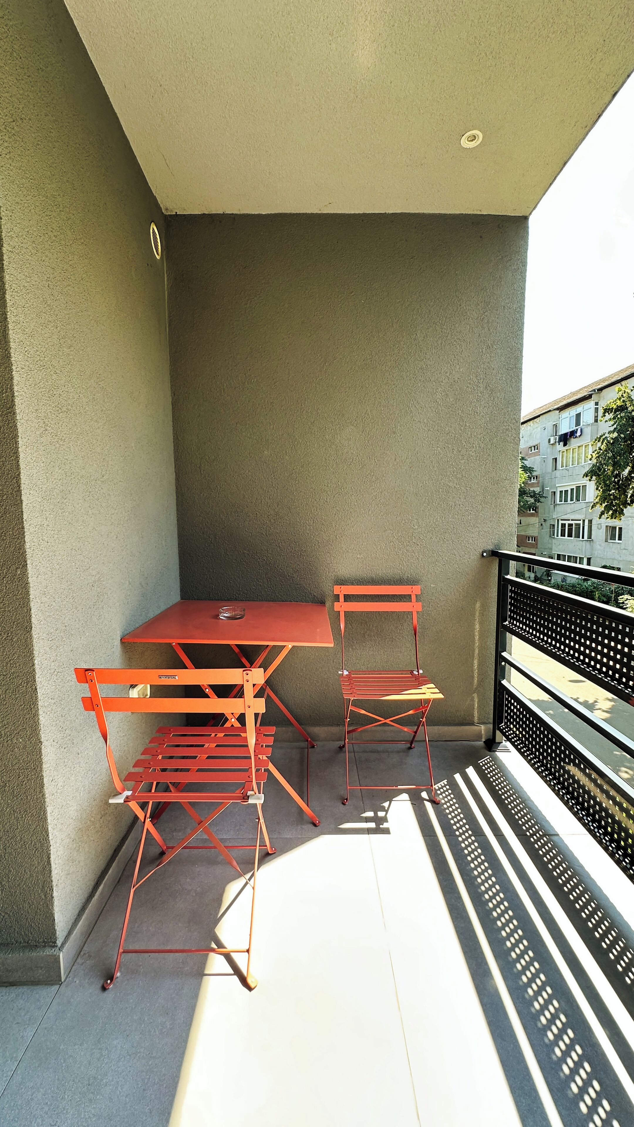 A balcony with a red metal table and chairs, a small window, and a view of buildings in the background.
