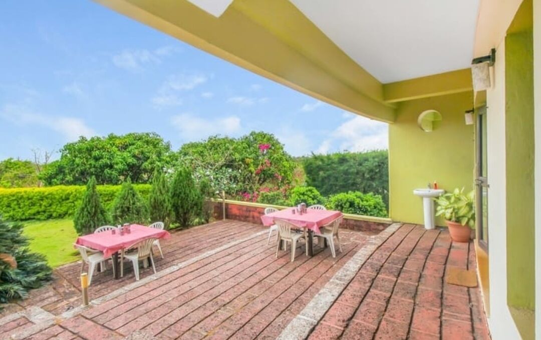Um pátio externo com duas mesas e cadeiras, um vaso de planta e vista para um jardim bem cuidado.