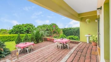Um pátio externo com duas mesas e cadeiras, um vaso de planta e vista para um jardim bem cuidado.