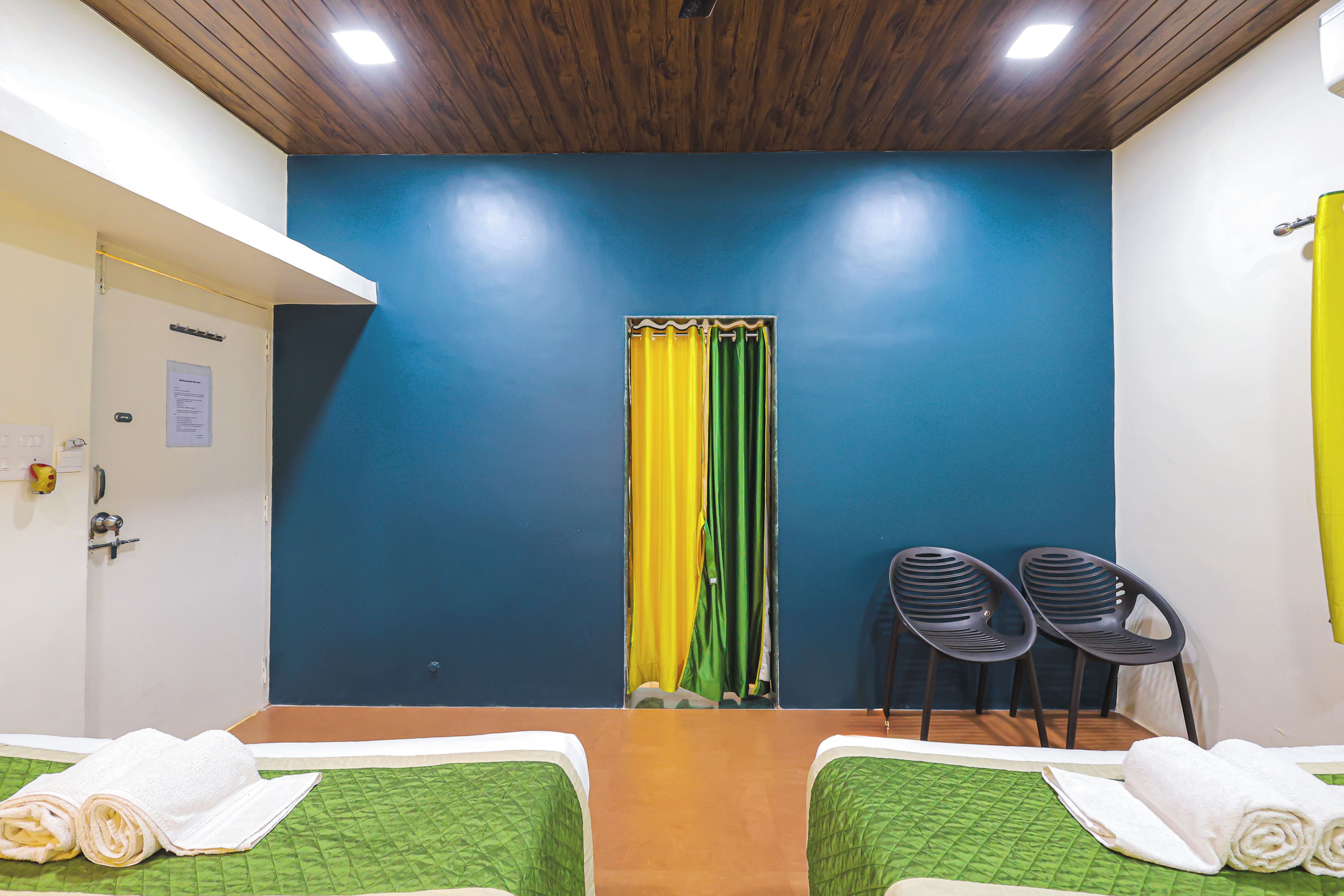 A hotel room with two beds, a wooden ceiling, a blue wall, a yellow curtain, and two black chairs.