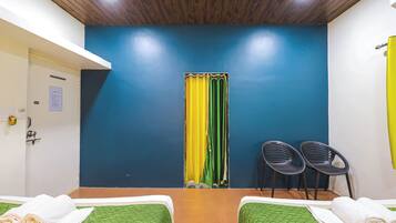 A hotel room with two beds, a wooden ceiling, a blue wall, a yellow curtain, and two black chairs.