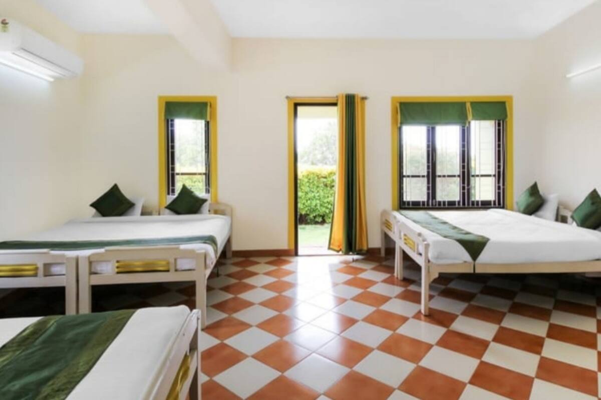 A room with two single beds, each with a green pillow and a white bedspread. There are two windows with yellow frames and green curtains. The floor has a checkered pattern.