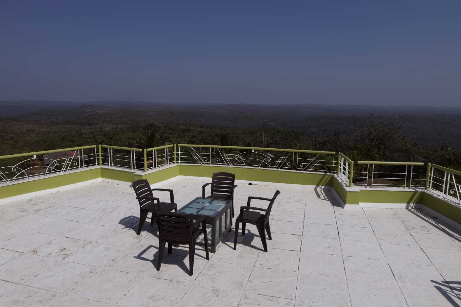A terrace with a table and chairs, surrounded by a green railing, overlooking a mountainous landscape.