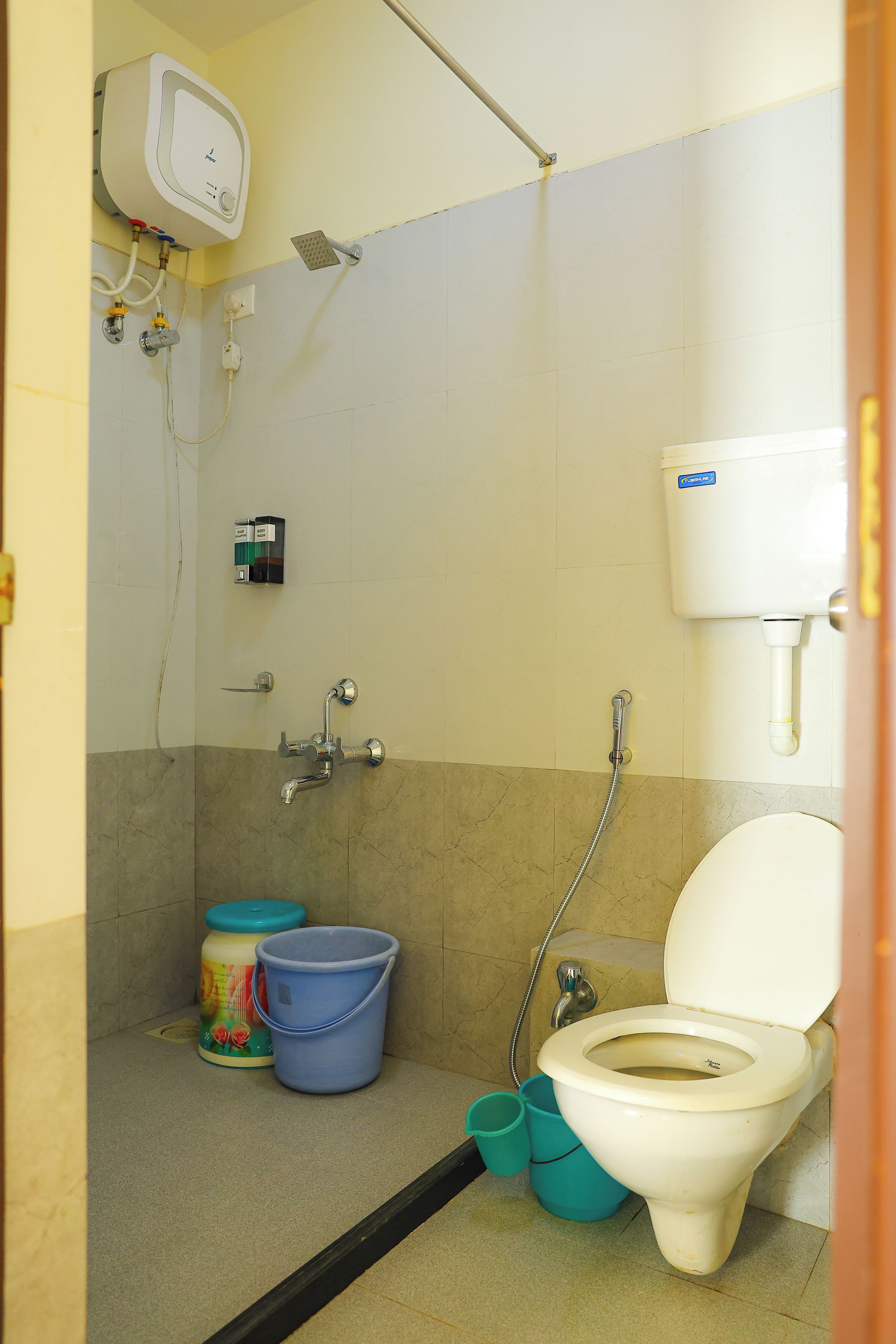 Une petite salle de bain compacte, équipée de toilettes, d’une douche et d’un lavabo. On y trouve deux seaux, un bleu et un vert, ainsi qu’un distributeur de savon fixé au mur.