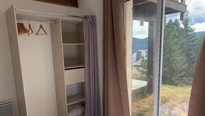 Een slaapkamer met houten vloer, een bed met een beddengoed met patroon, een open kledingkast, en uitzicht op een bergachtig landschap door een groot raam.