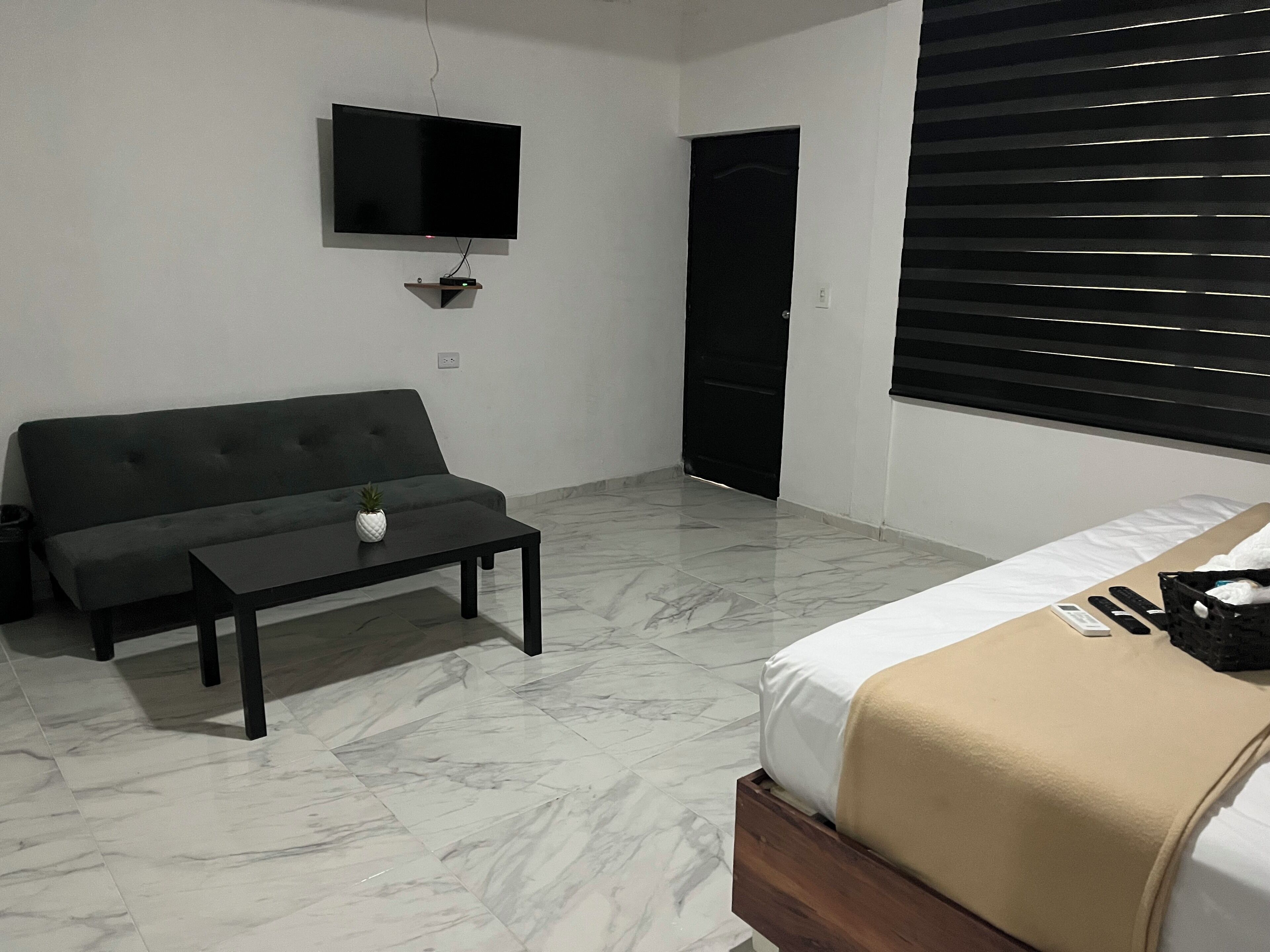 A modern room with a sofa, a small table with a plant, a bed with a beige blanket, and a TV mounted on the wall.