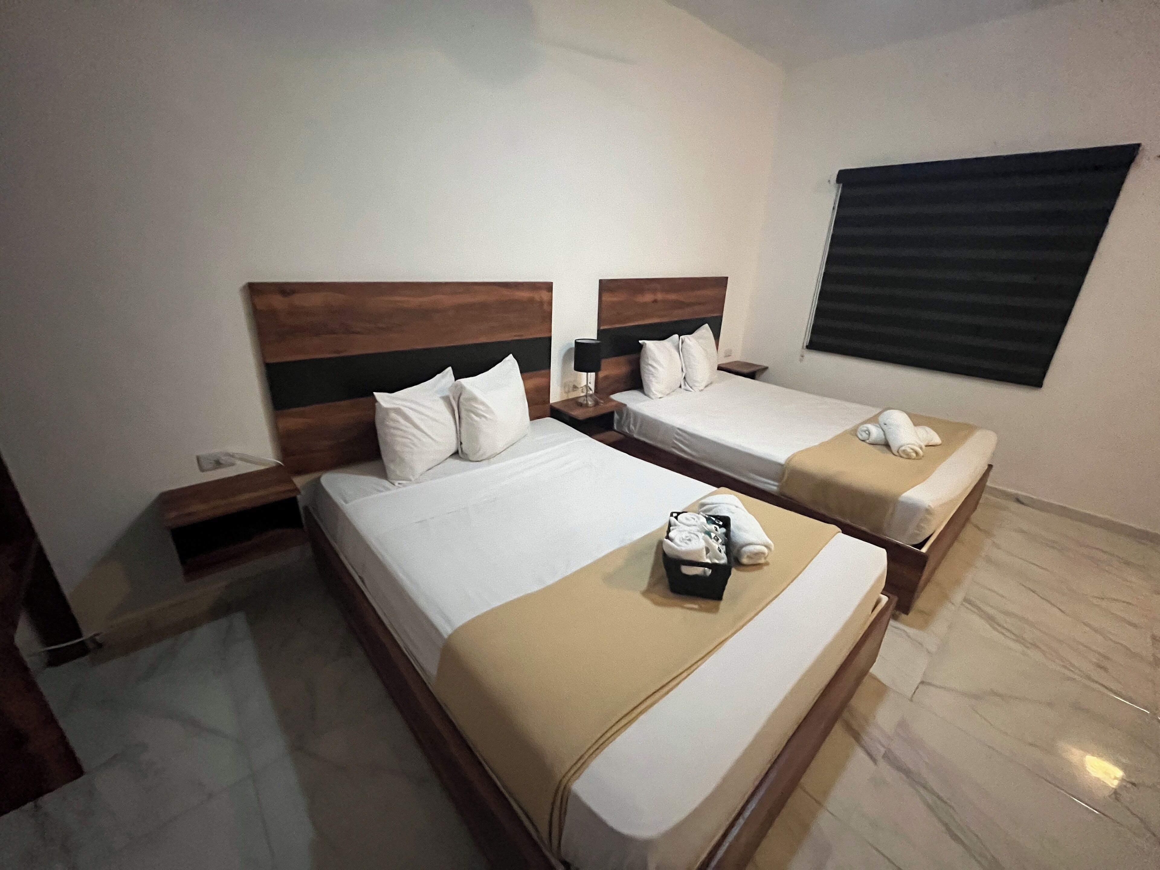 Een hotelkamer met twee bedden, elk voorzien van houten hoofdborden en witte beddengoed. Er staat een klein tafeltje met een mand handdoeken en een klok aan de muur.