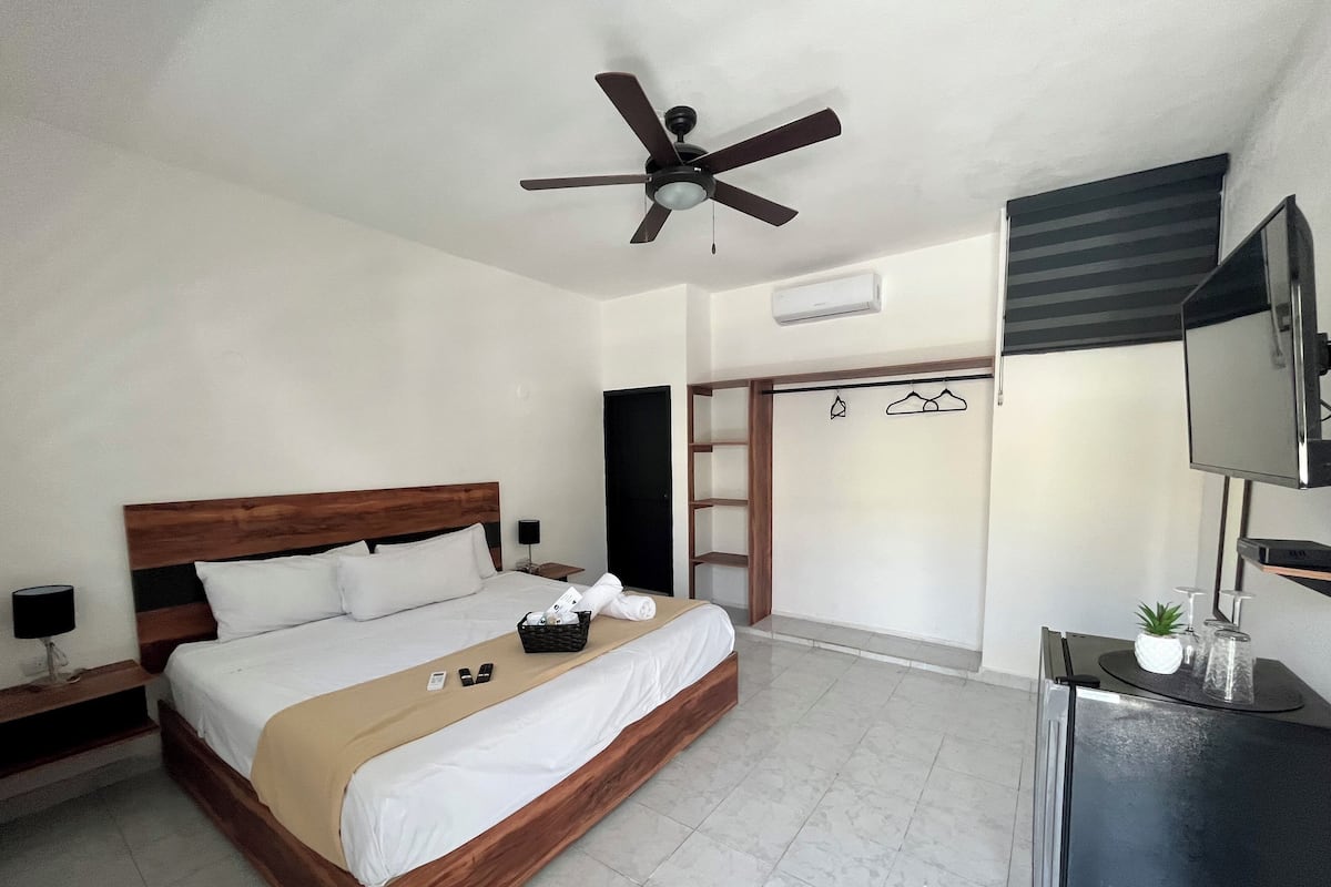 Quarto de hotel com cama de madeira, ventoinha no teto, televisão, guarda-roupa e uma pequena mesa com uma planta.