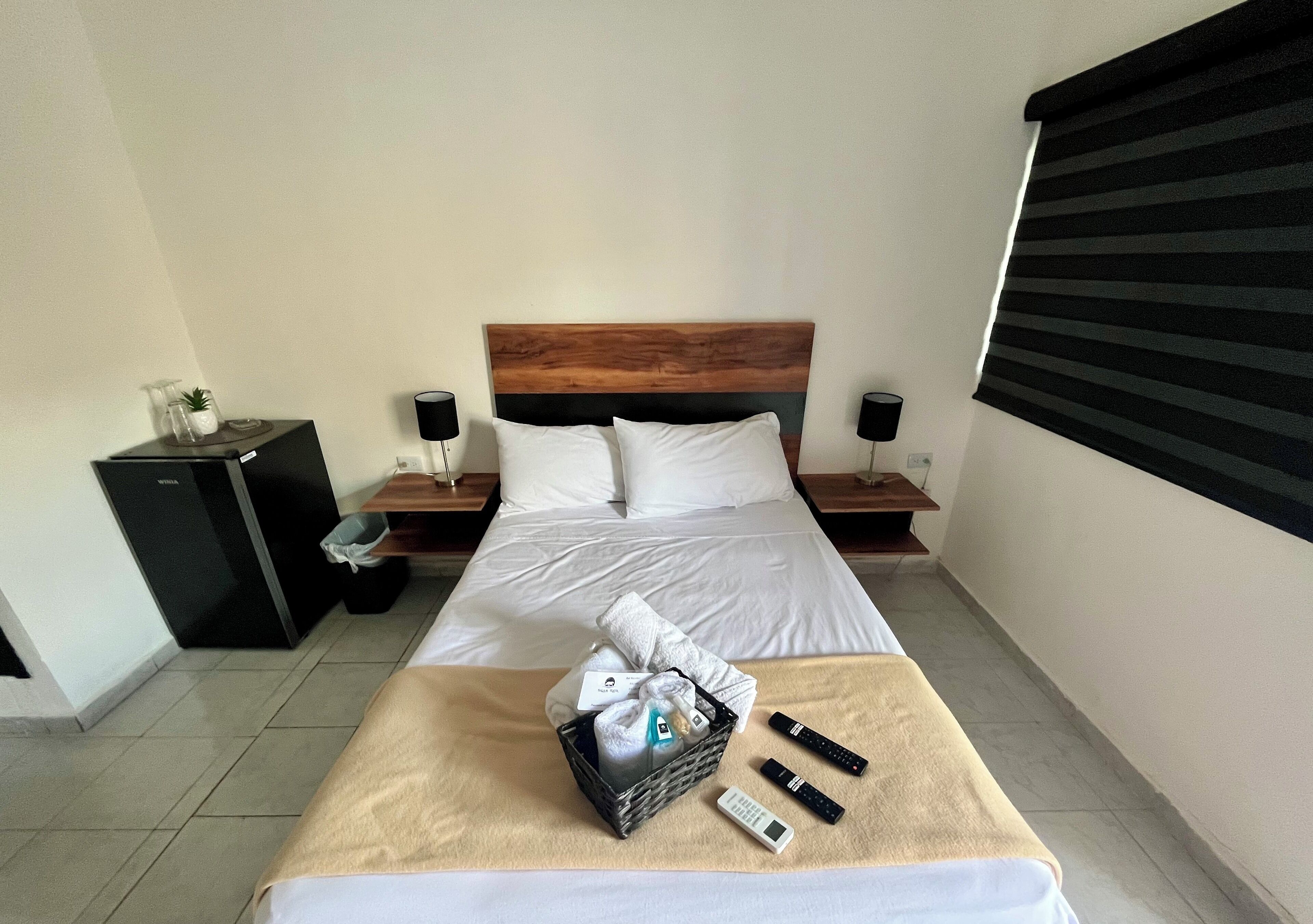 A hotel room with a bed, bedside tables, a small desk, a lamp, a window with blinds, and a basket with toiletries.