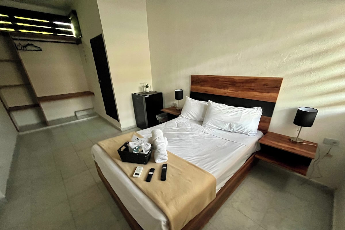 Quarto de hotel com cabeceira de madeira, cama com roupa de cama branca, mesa de cabeceira com abajur e um pequeno alto-falante preto.