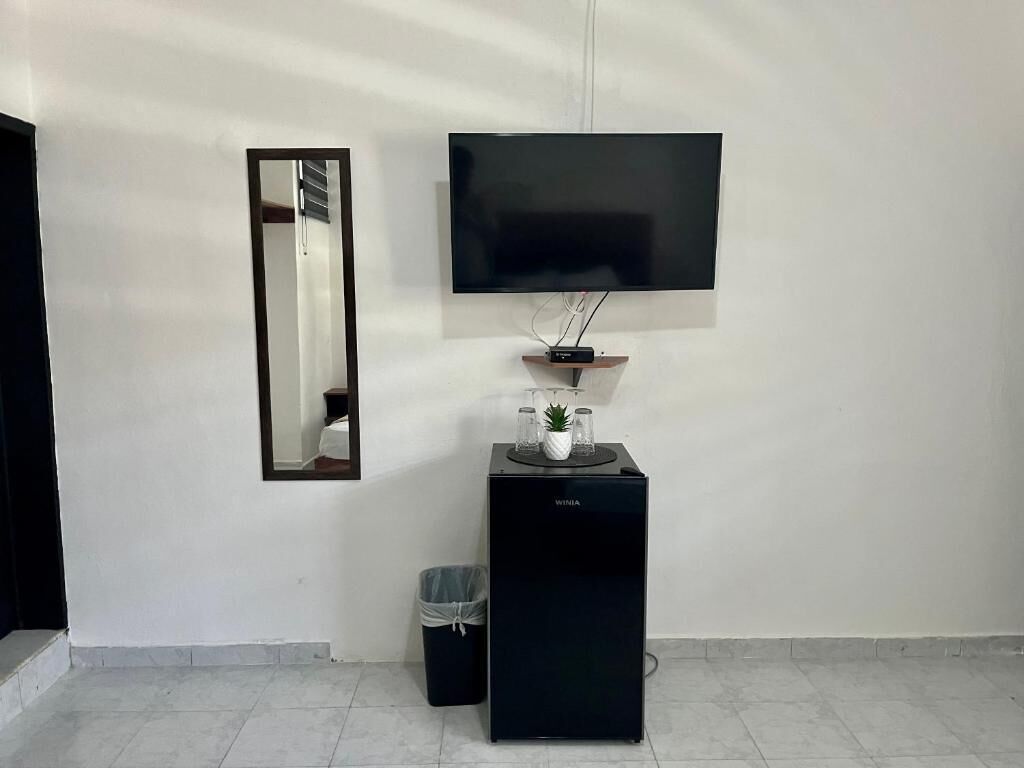 Una habitación con una televisión de pantalla plana montada en la pared, un altavoz negro pequeño debajo, y un bote de basura a la izquierda.