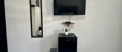 Una habitación con una televisión de pantalla plana montada en la pared, un altavoz negro pequeño debajo, y un bote de basura a la izquierda.