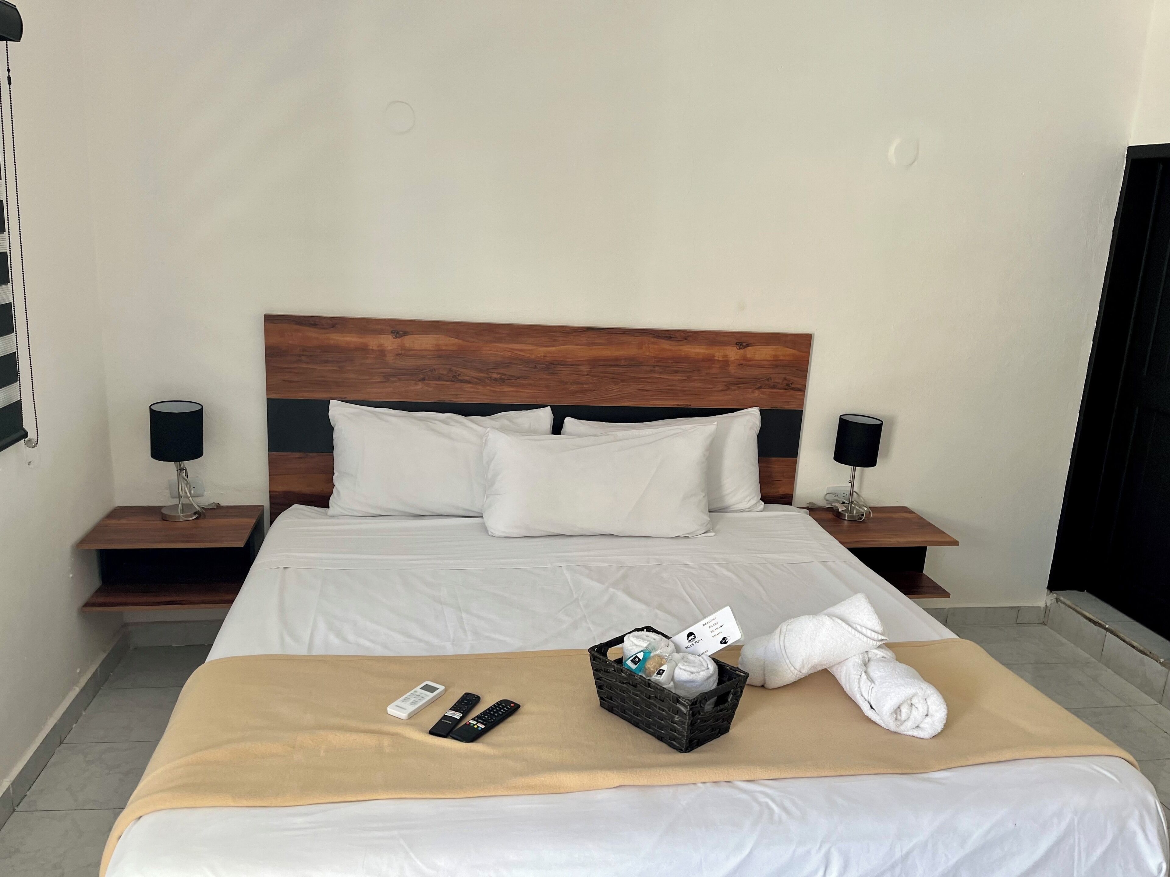 Un lit bien fait, avec du linge de lit blanc, un plaid beige et un panier rempli d’articles de toilette.