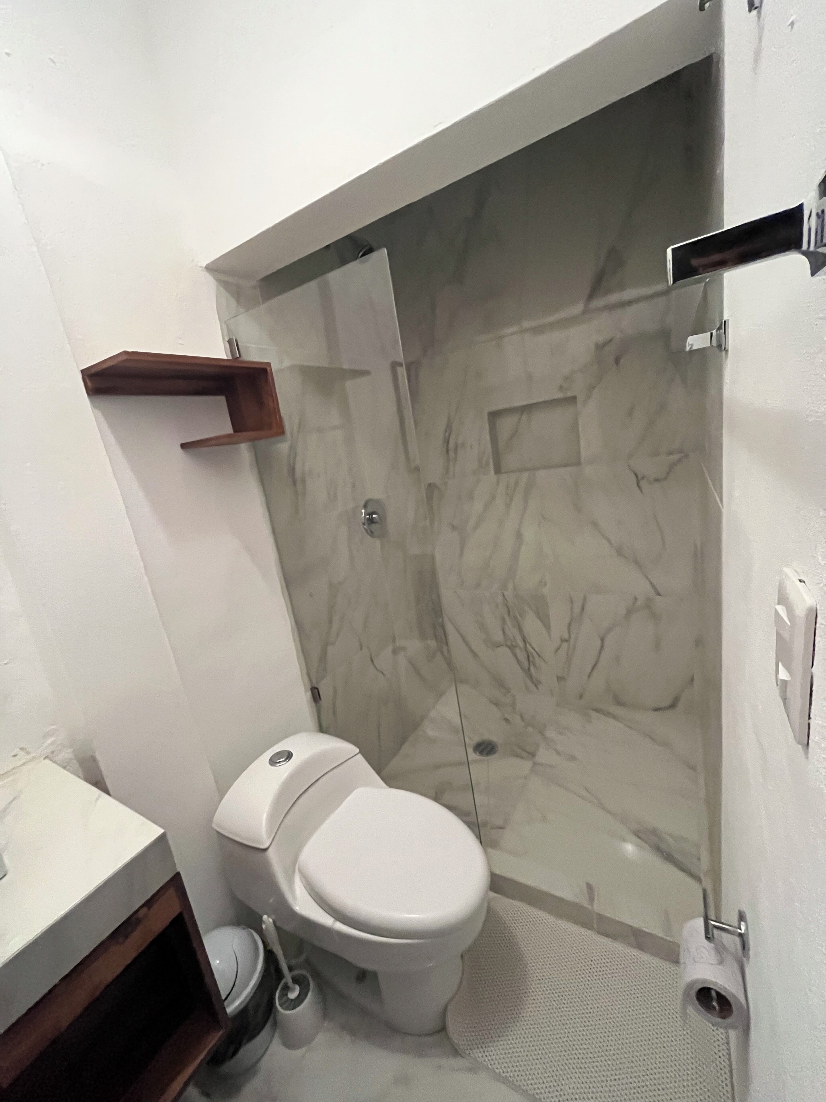 Une salle de bain comprenant des toilettes, un espace douche, un mur recouvert de carreaux de marbre et une étagère en bois.