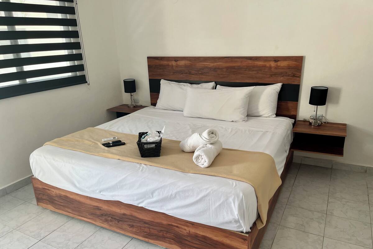 Cama bem arrumada com cabeceira de madeira, roupa de cama branca e manta bege. Há dois controles remotos e um cesto com toalhas na cama.