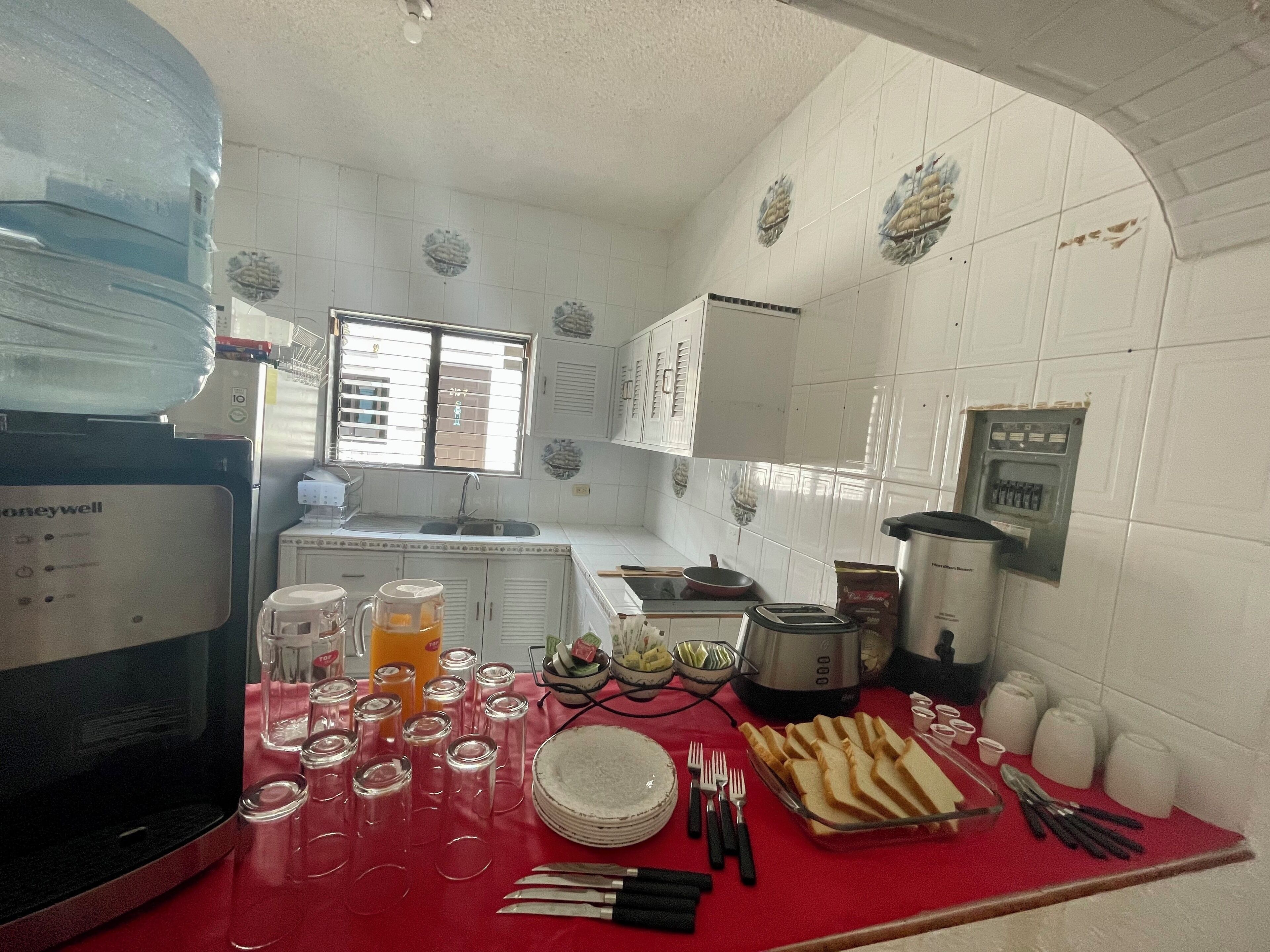 Una cocina con paredes de azulejos blancos, una ventana con persianas y diversos electrodomésticos y utensilios de cocina sobre la encimera.
