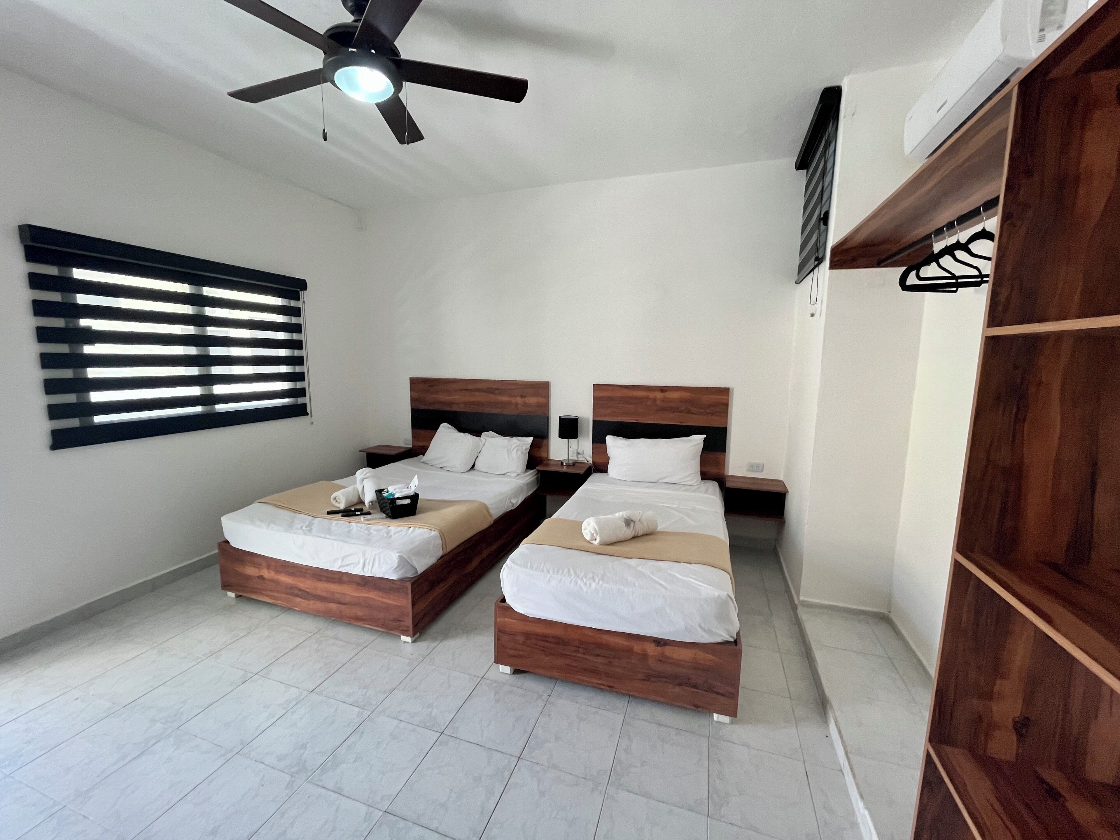 A hotel room with two wooden beds, a ceiling fan, a window with blinds, and a wardrobe.