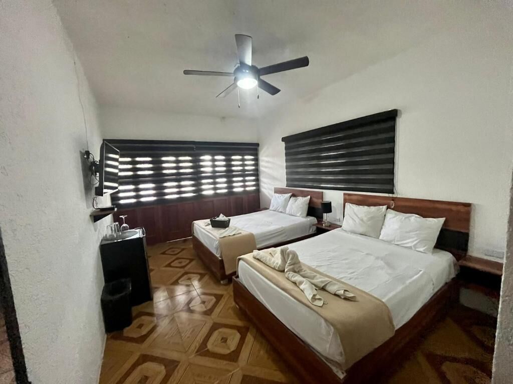 A hotel room with two beds, a ceiling fan, a window with blinds, a small table, and a trash bin.