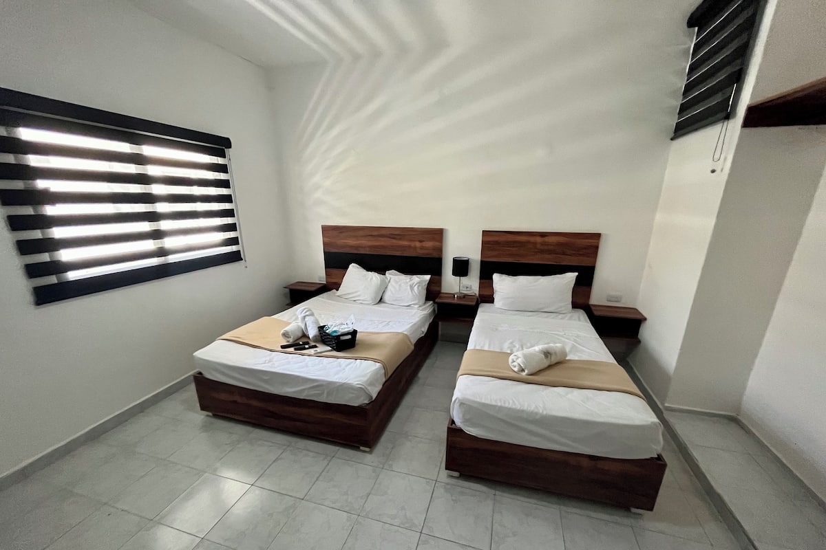 Quarto de hotel com duas camas, uma janela grande com persianas, mesa de cabeceira com abajur e piso ladrilhado.