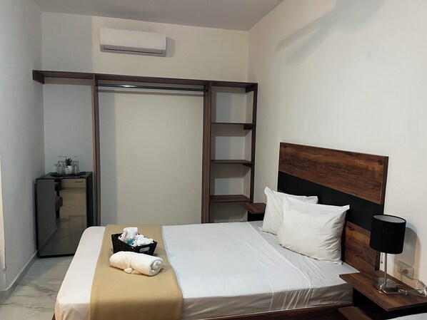 A hotel room with a bed, a nightstand, a lamp, a wooden headboard, a towel, a basket, and a wardrobe with a mirror.