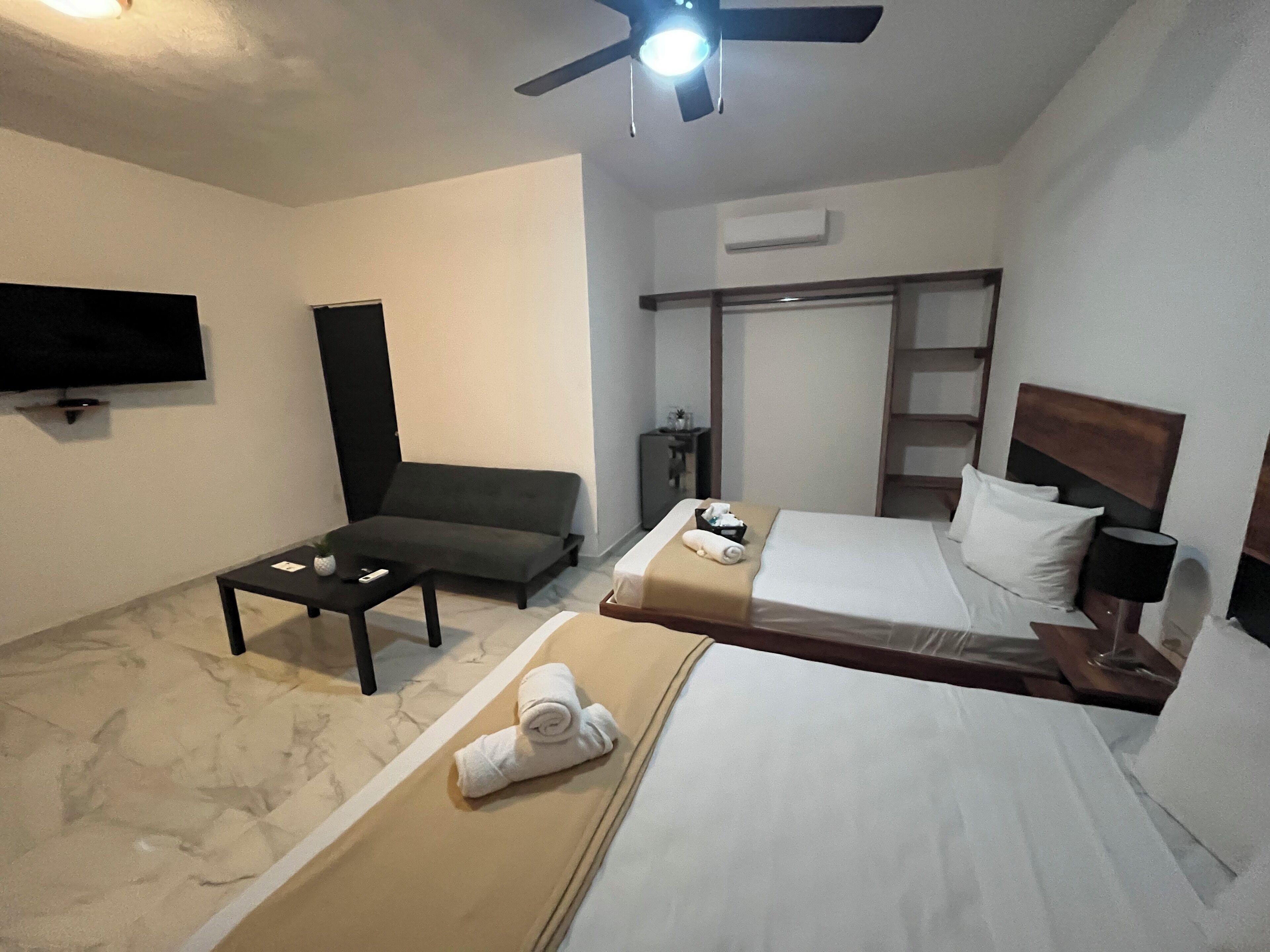 A hotel room with a bed, a sofa, a TV, a ceiling fan, and a small table with a remote control.