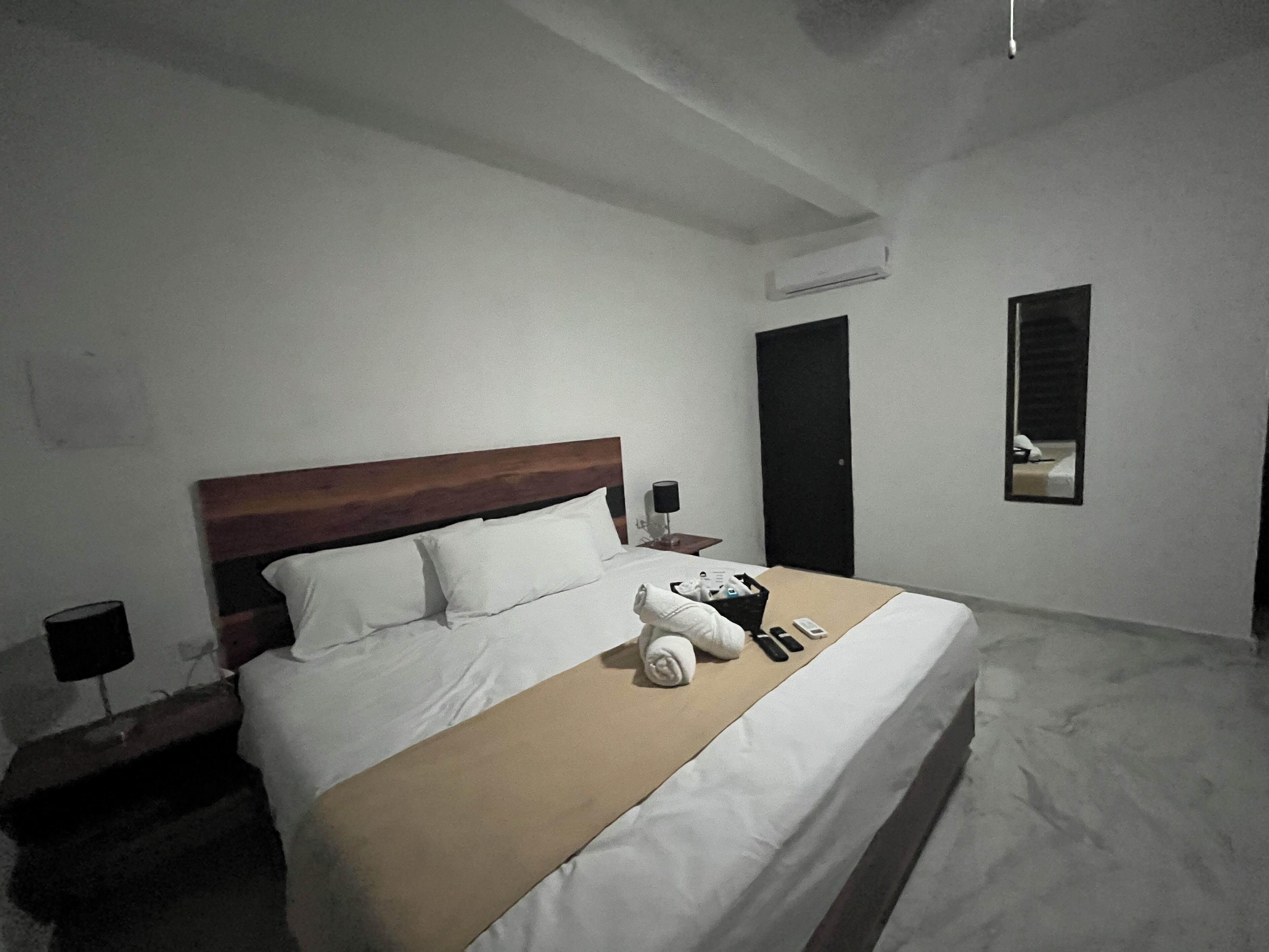 A neatly made bed with white linens and a wooden headboard. There is a small bedside table with a lamp and a remote control. A mirror is mounted on the wall to the right of the bed.