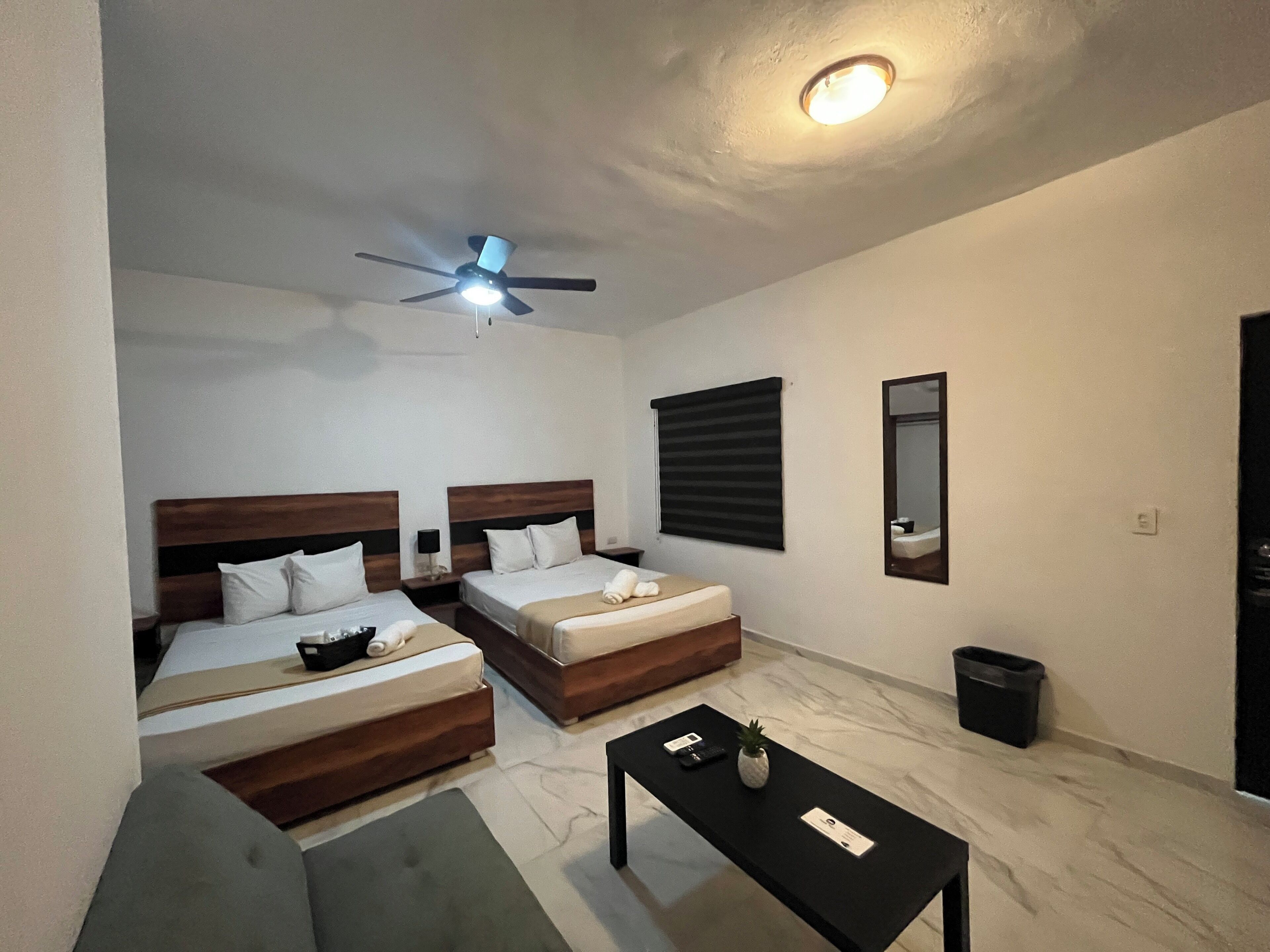 A hotel room with two beds, a ceiling fan, a small table, a trash bin, and a mirror.