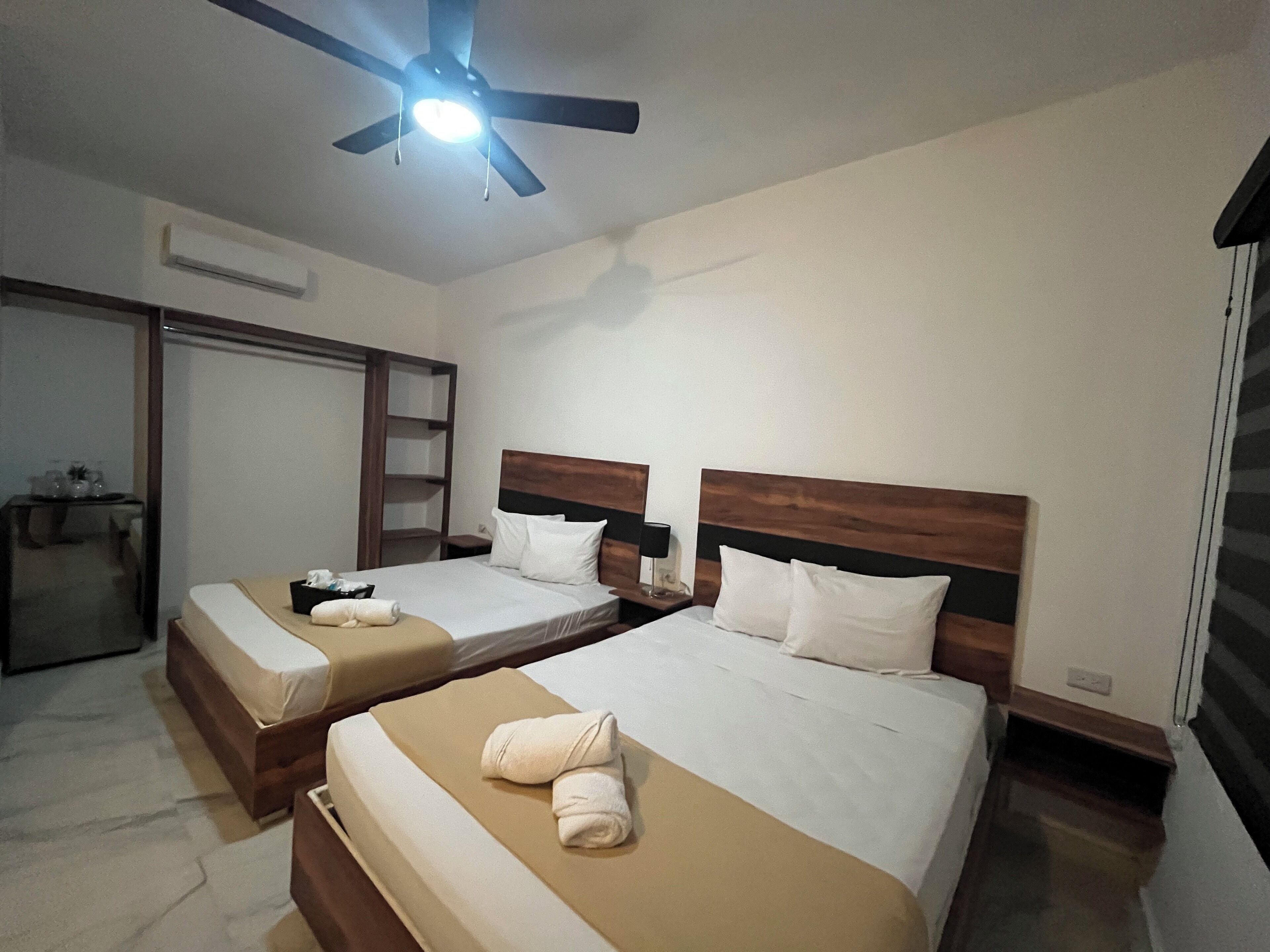 Een hotelkamer met twee bedden, een plafondventilator, een kledingkast en een nachtkastje met een lamp.