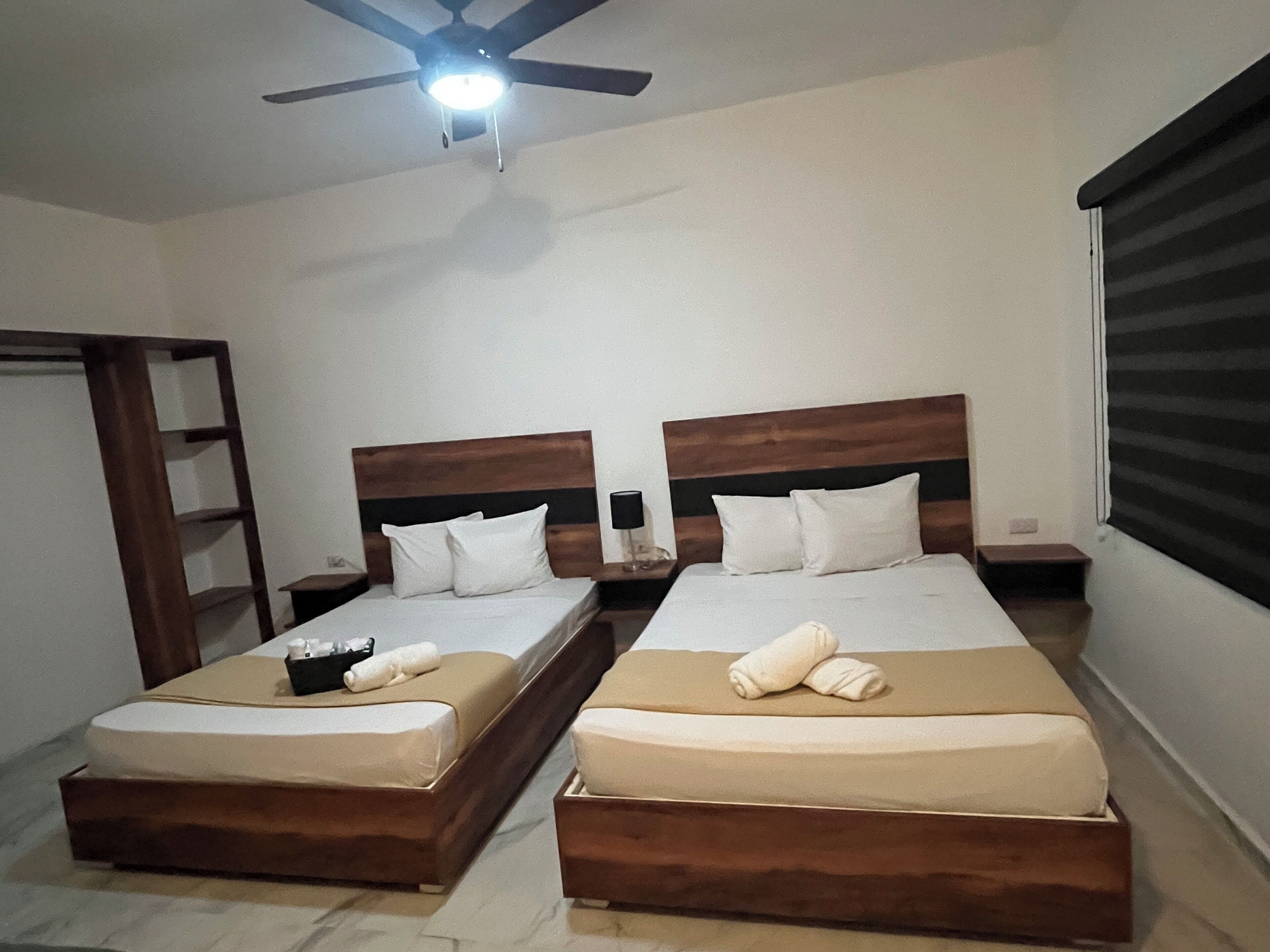 Habitación de hotel con dos camas, cabeceros de madera, ropa de cama blanca, ventilador de techo y una ventana con persianas.