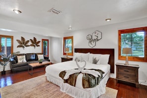 A spacious bedroom with a large bed, a dark leather sofa, a wooden nightstand, and a window with a view of greenery.