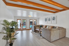 A spacious living room with a beige sectional sofa, a coffee table, and a large potted plant. There are framed botanical prints on the wall and a view of a pool through glass doors.