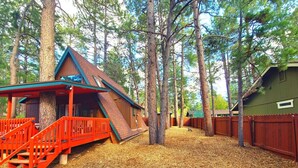 Une cabane avec un escalier rouge et un toit en pente, entourée de grands arbres et dotée d’une terrasse en bois.