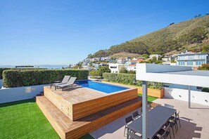 Una piscina en la azotea con mobiliario de terraza de madera y vistas a un pueblo en la colina.