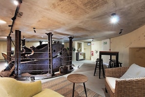 Una habitación con una máquina industrial grande, un sofá de mimbre, una mesita redonda y una zona de bar.