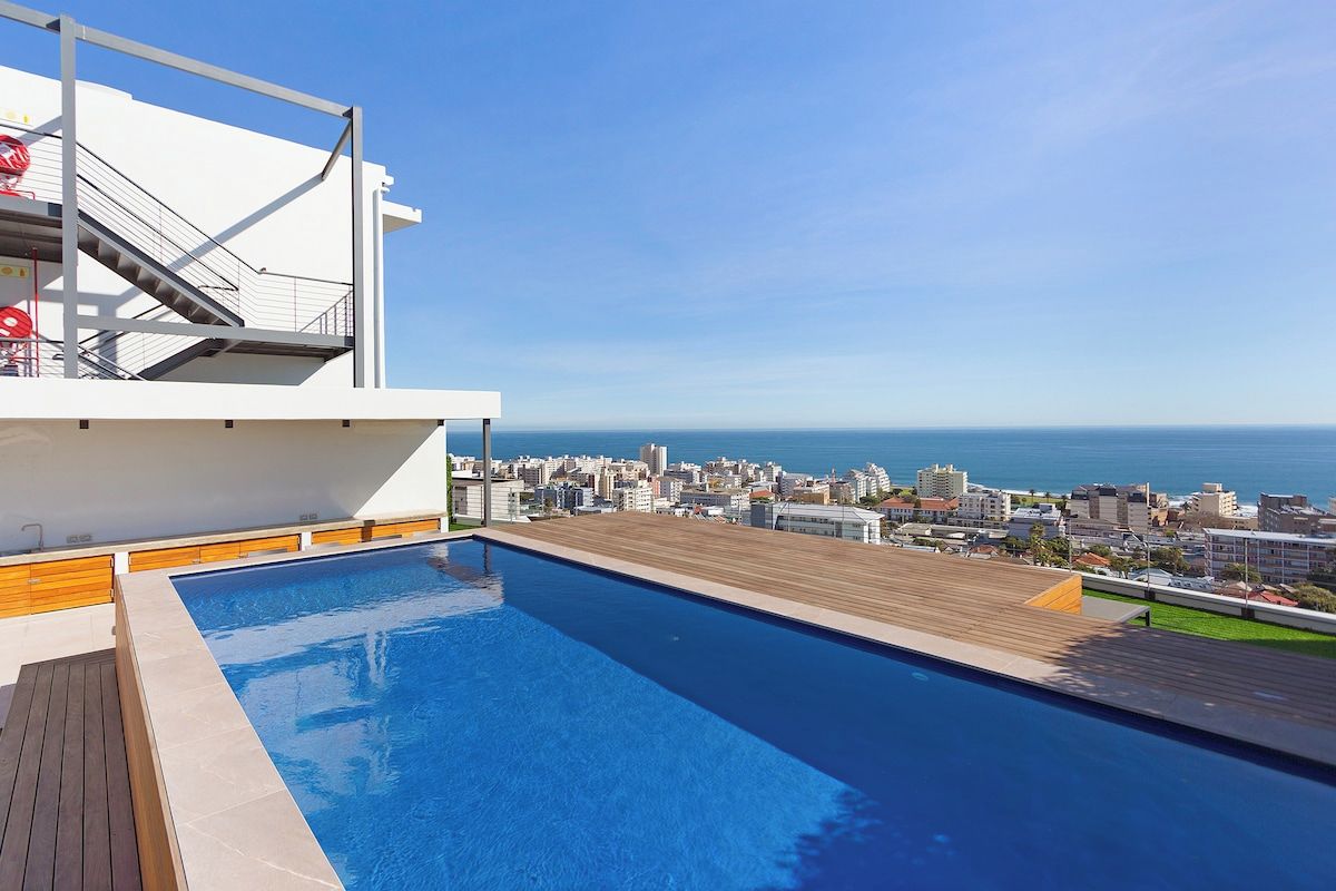 A rooftop swimming pool with a city view, a wooden deck, and a white building structure.