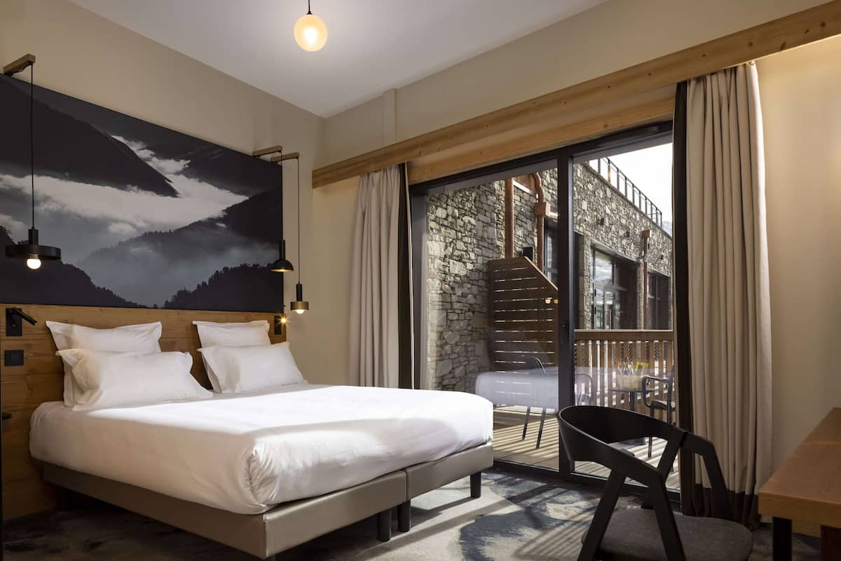 Une chambre comprenant un lit, une chaise, une table, une grande fenêtre et une vue sur un escalier en pierre menant à un espace extérieur.