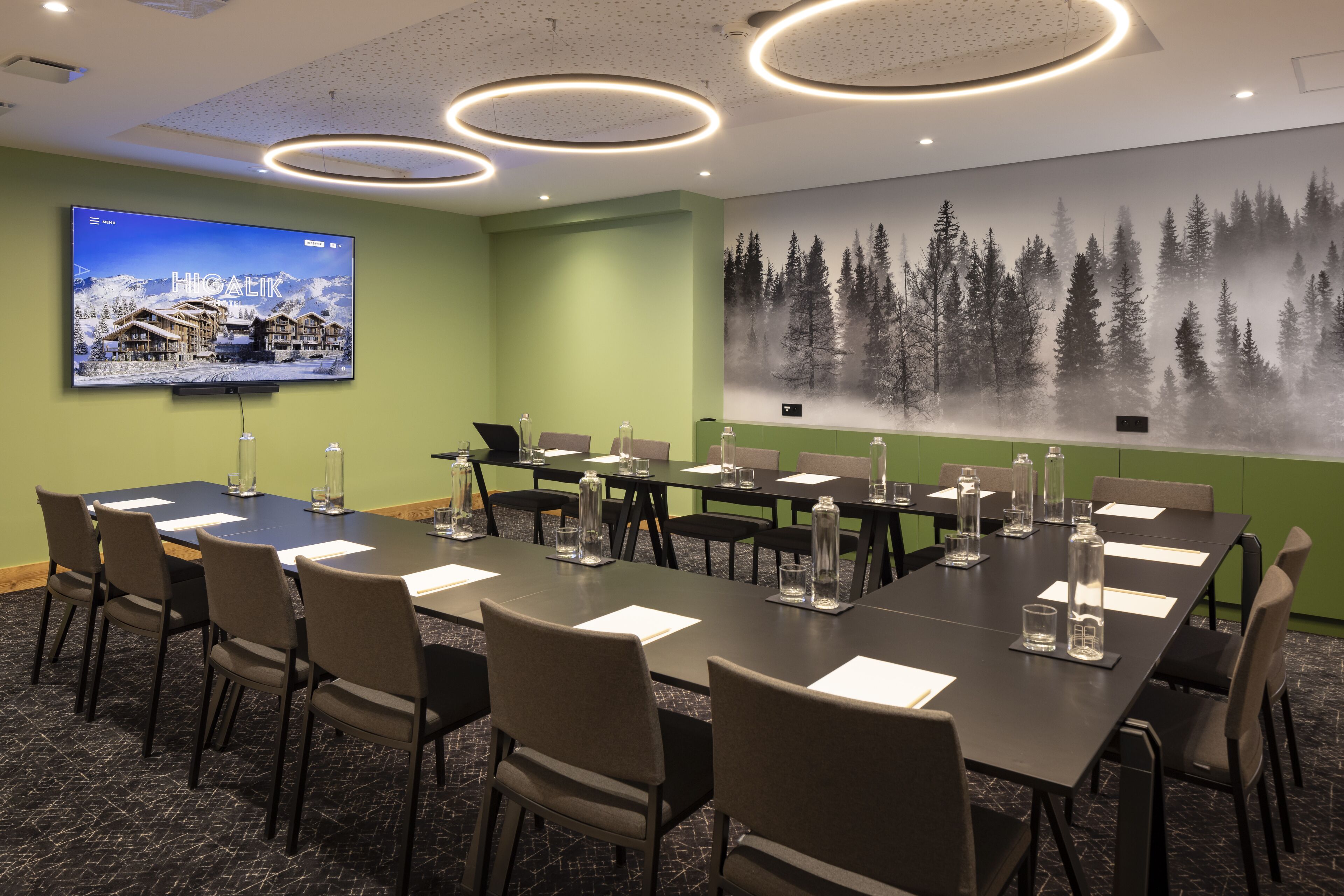 A conference room with a large flat-screen TV displaying a snowy mountain resort, green walls, a forest mural, and a long table with chairs and water bottles.