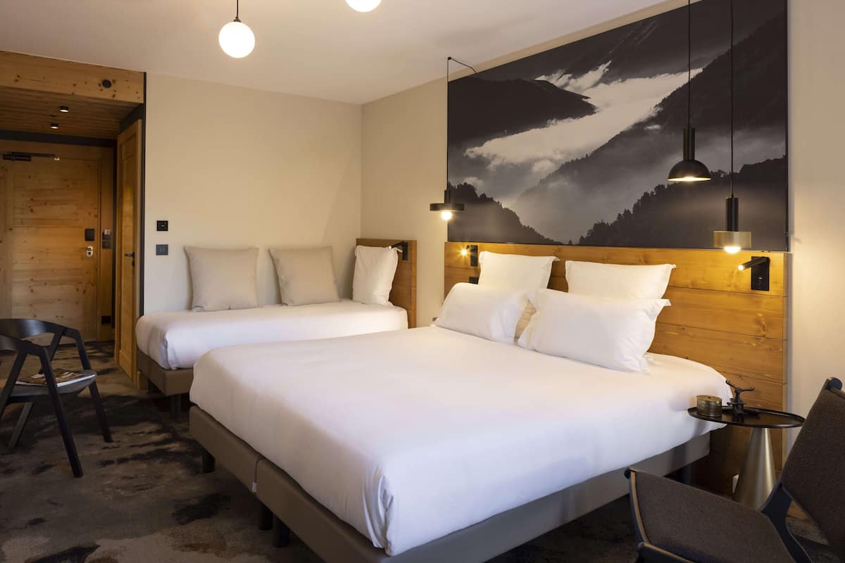 Une chambre d’hôtel avec deux lits, une tête de lit en bois, un grand tableau représentant un paysage de montagne et une petite table ronde avec une chaise.