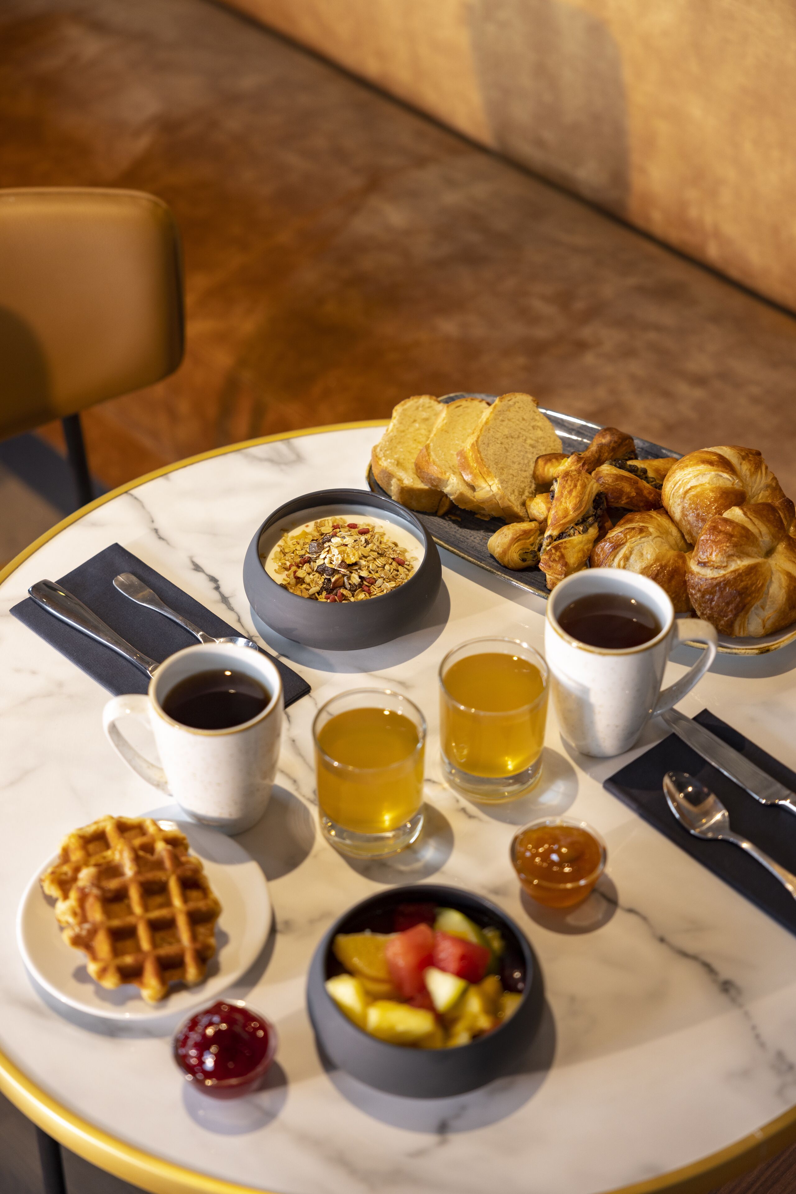 A breakfast table with a waffle, fruit bowl, croissants, and various beverages.