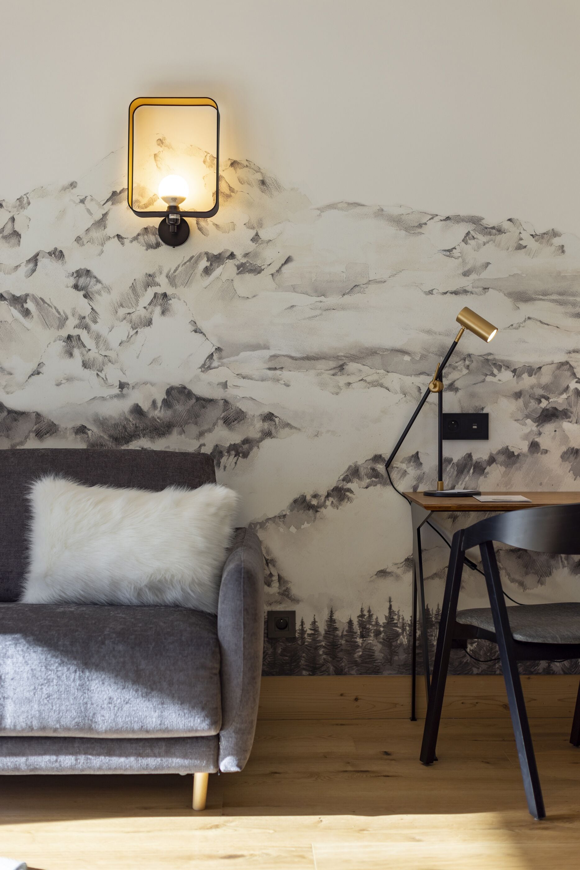 Ett rum med en grå soffa, en vit, fluffig kudde, ett skrivbord med en stol, en vägglampa och en vägg prydd med en väggmålning som föreställer ett berg.