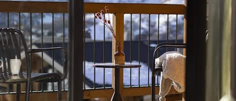 A wooden deck with a view of snow-covered mountains. There are two chairs and a small table with a vase of dried flowers.