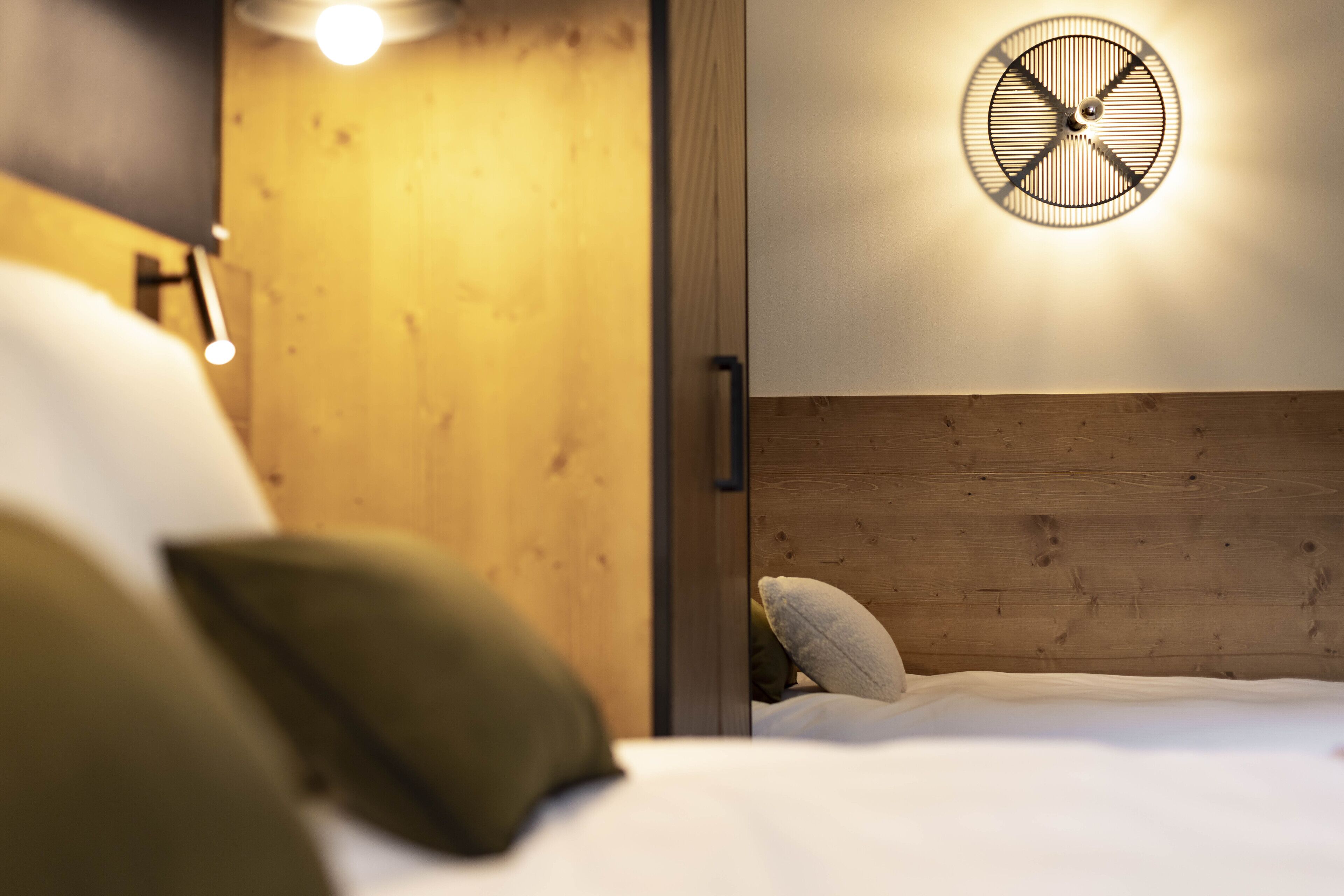 Un dormitorio con cabecera de madera, lámpara redonda fijada a la pared y cama con ropa blanca y almohadas.