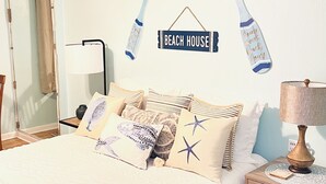 Un dormitorio con cama, almohadas, una mesita de noche, lámpara y decoración de pared que incluye un letrero que dice 'Beach House' y un murciélago.