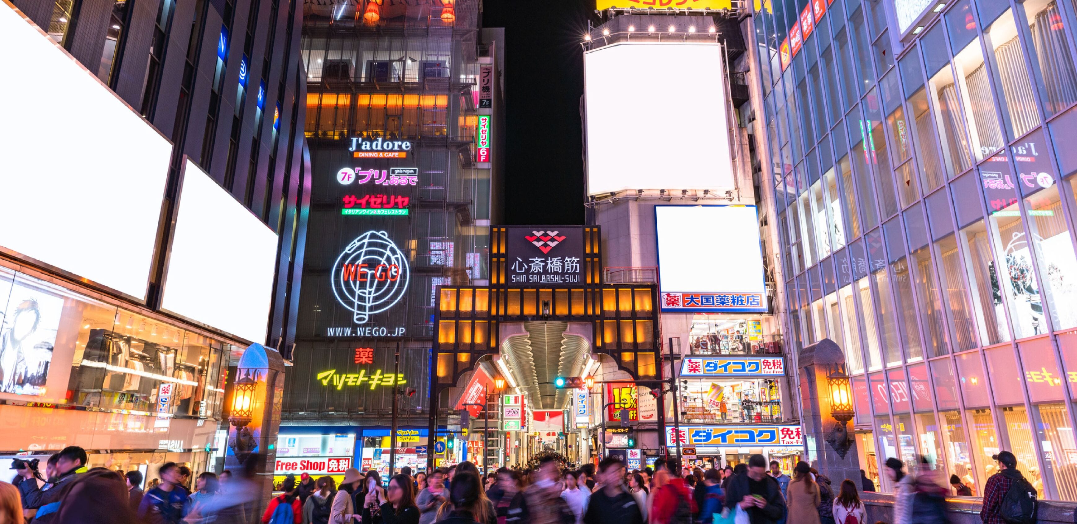 Une rue animée d’une ville, de nuit, avec des enseignes et des panneaux publicitaires illuminés, dont une grande enseigne « WEGO » bien visible.