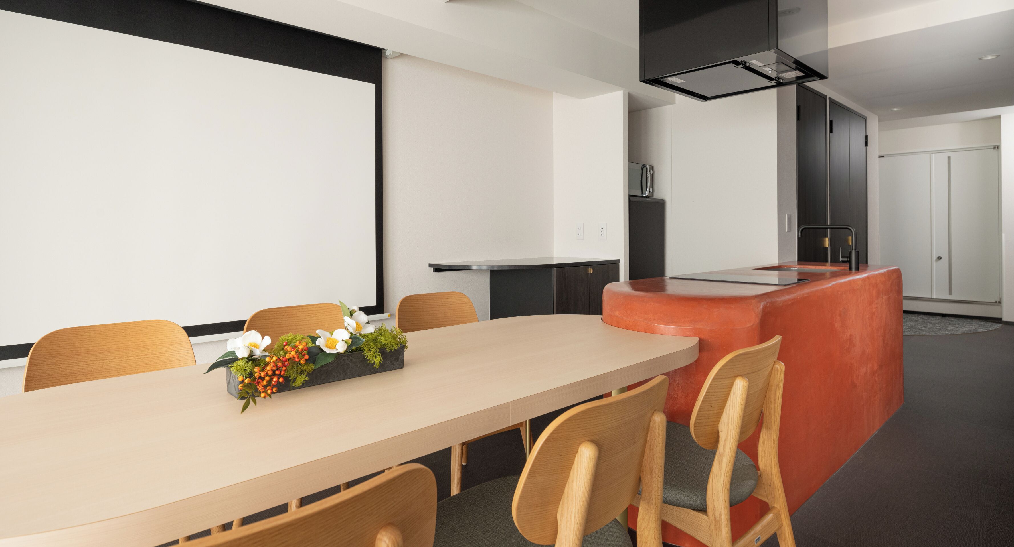 A modern conference room with a long table, wooden chairs, a small plant, and a projector screen.