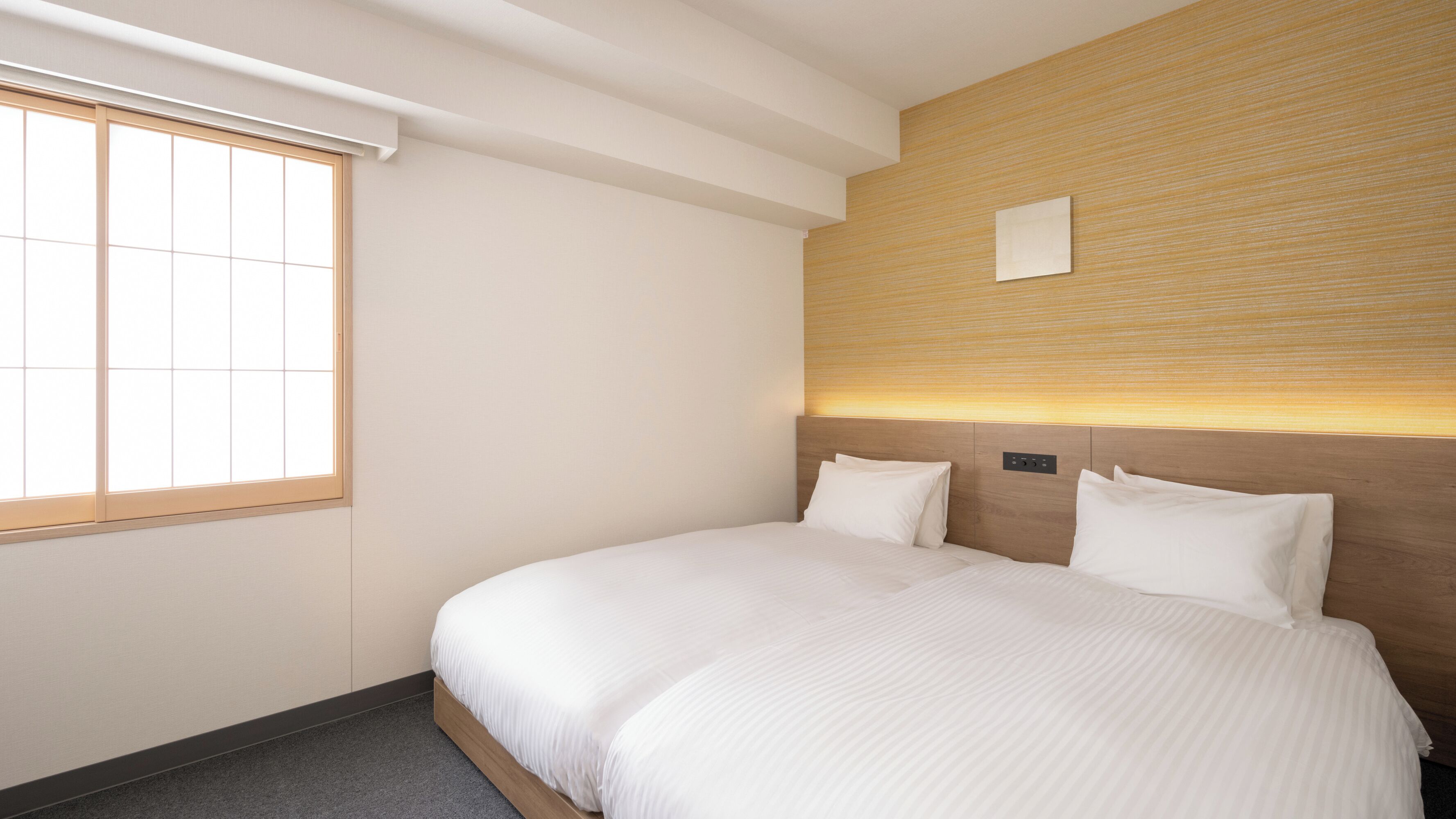A hotel room with a large bed, a window with blinds, a wooden headboard, and a wall-mounted light fixture.