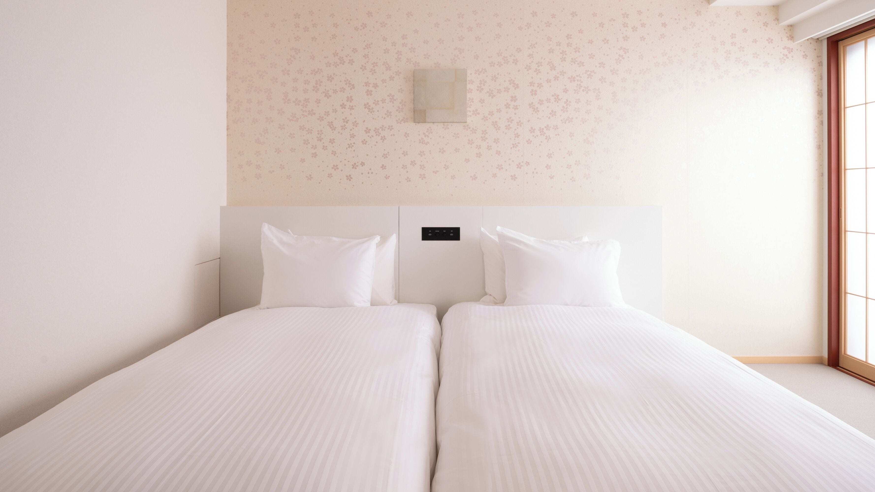 A double bed with white linens in a room with a floral wallpaper pattern and a small wall-mounted light fixture.