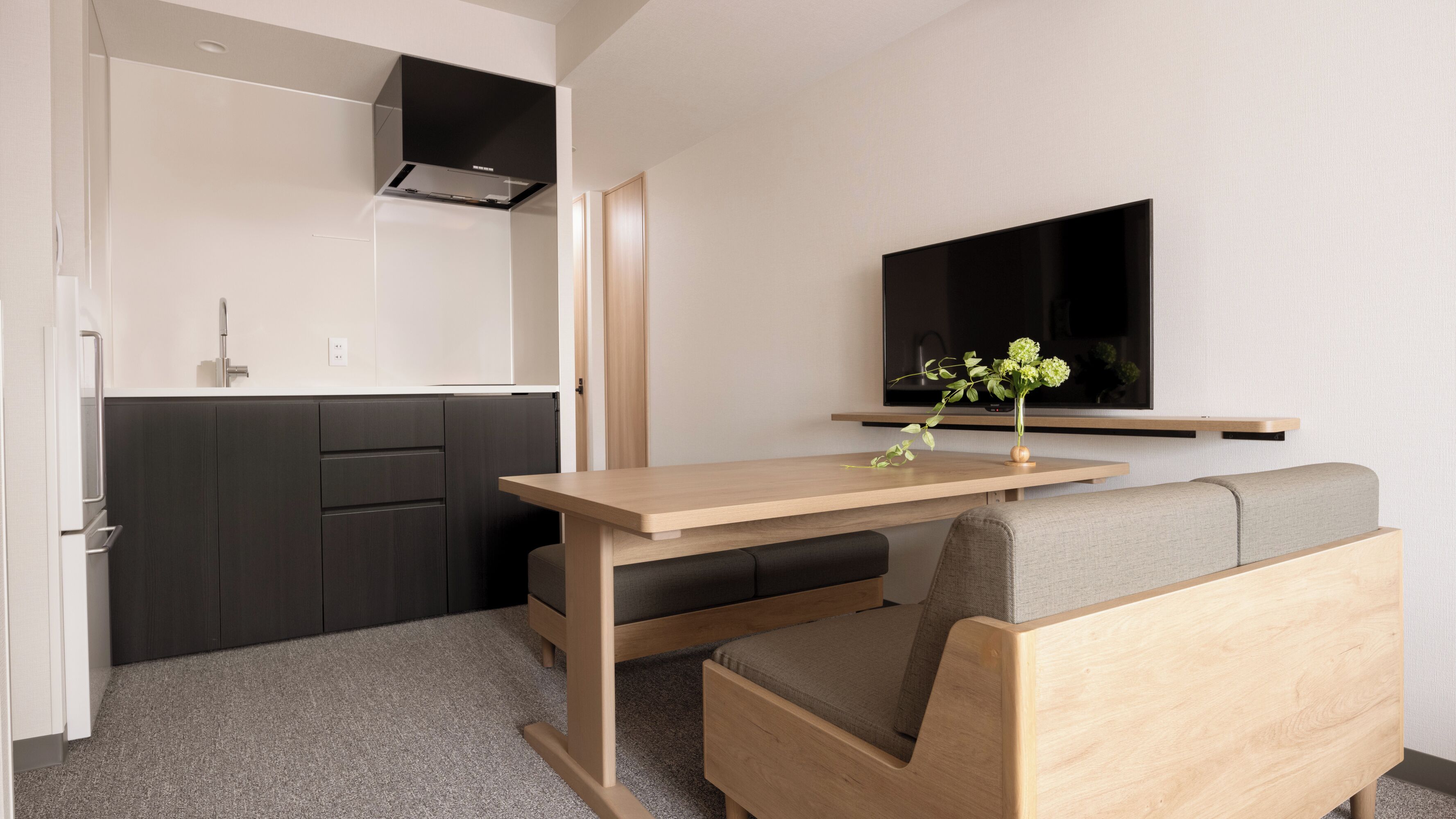 A modern kitchen with dark cabinets, a wooden dining table, and a grey sofa. A flat-screen TV is mounted on the wall above the sofa.