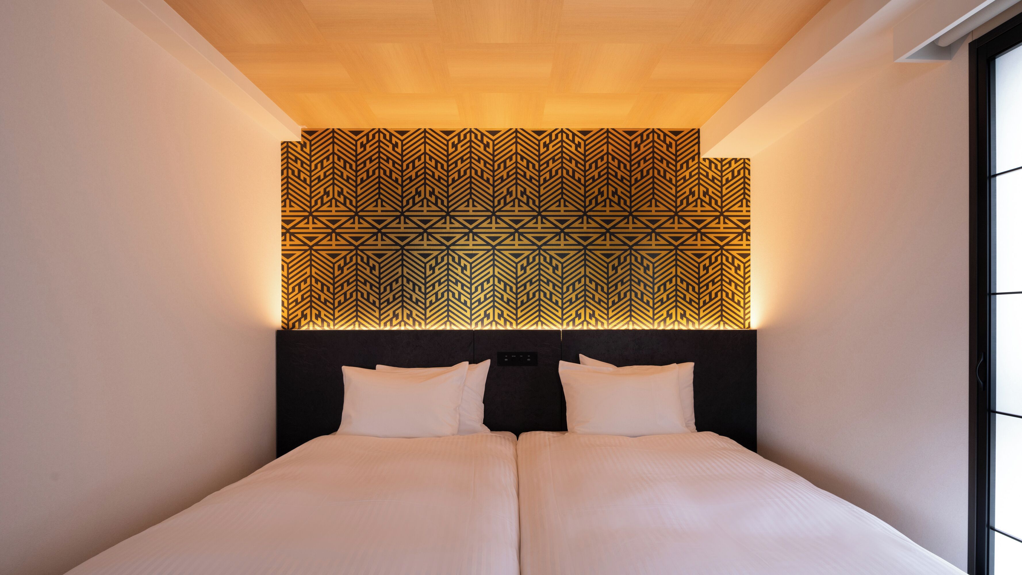 Une chambre d’hôtel moderne avec une tête de lit à motifs géométriques, un grand lit avec du linge de lit blanc et une vue sur le ciel par la fenêtre.