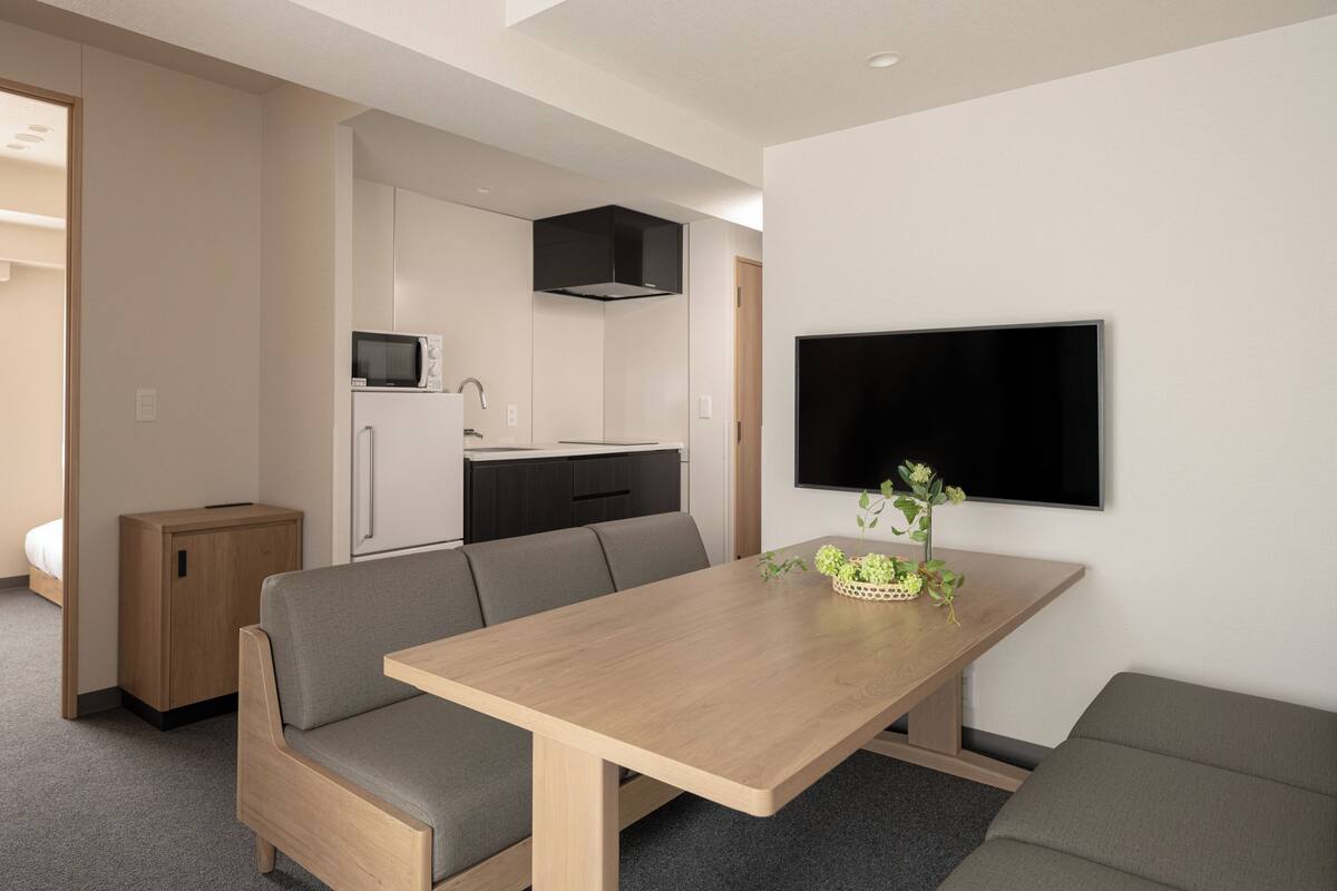 Un espacio habitable compacto con kitchenette, una zona de comedor con mesa y dos sofás, un televisor montado en la pared y un mueble pequeño.